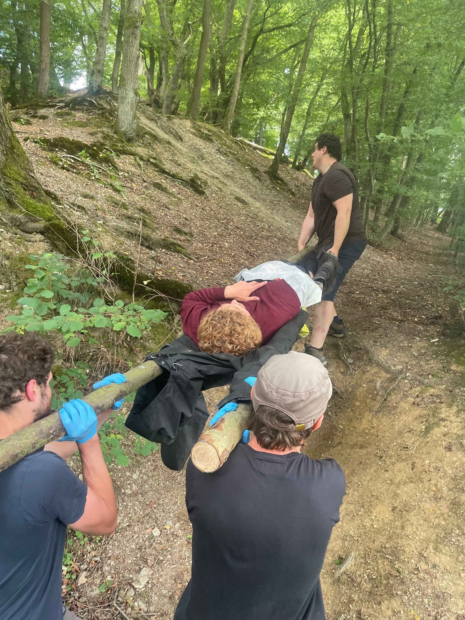 Eine Gruppe trägt eine verletzte Person auf einer selbstgebauten Trage aus Baumstämmen im Wald