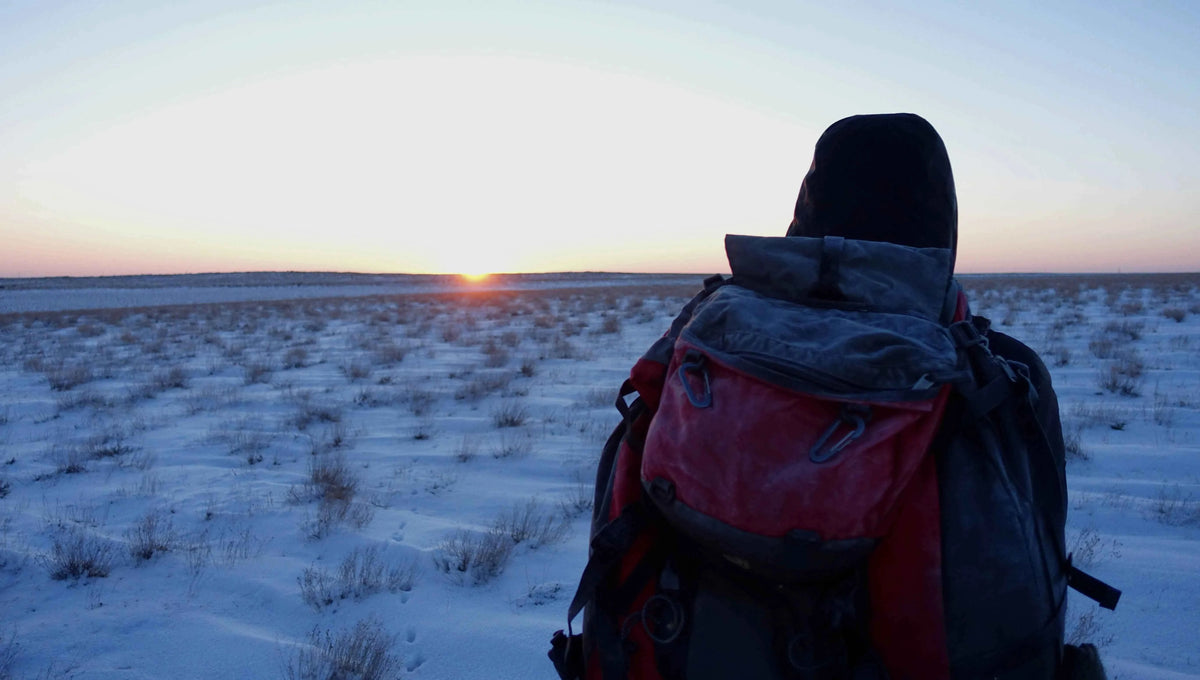 Person mit Rucksack blickt von hinten bei Sonnenaufgang über eine weite, verschneite Landschaft, Kasachstan
