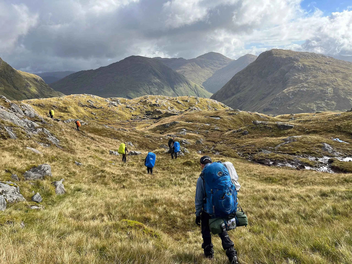 Eine Gruppe Wandernde mit großen Rucksäcken geht auf einem schmalen Pfad durch eine weite, bergige Graslandschaft.