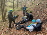 Praktische Übung im Wald: Ersthelfer versorgen eine am Boden liegende Person auf einem laubbedeckten Hangweg, während im Hintergrund ein Notruf per Funk abgesetzt wird, im Rahmen eines WFA 1 Essential Kurses